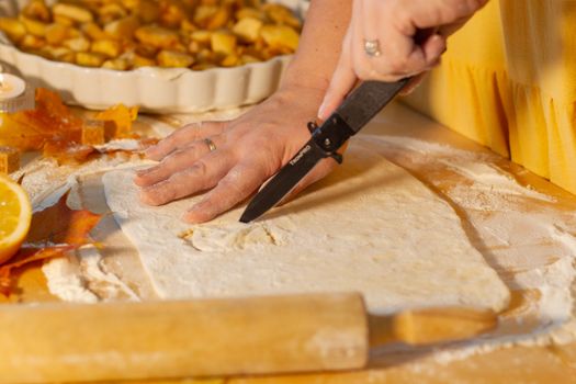 Main coupant la pâte à tarte pour une tarte aux pommes sur un plan de travail fariné, avec des pommes en arrière-plan.